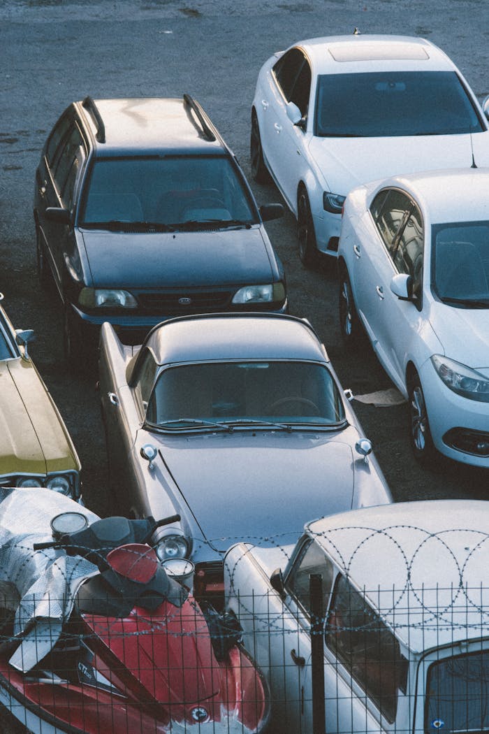A mix of classic and modern cars parked in an outdoor lot behind a barbed wire fence.